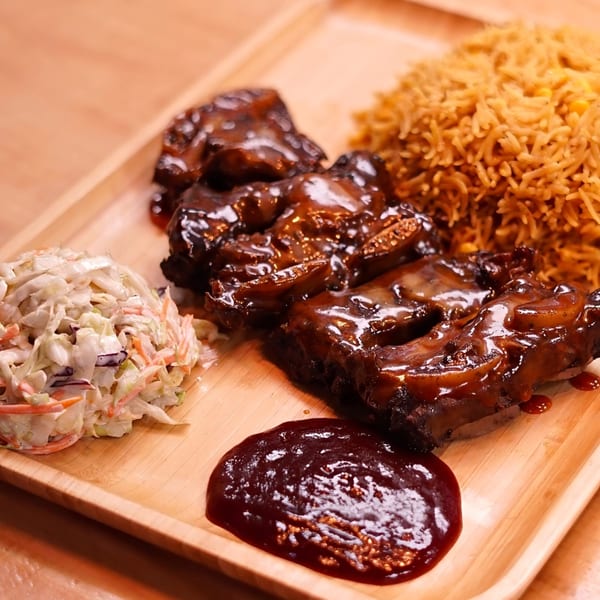 Peri-Peri ribs with rich sauce, golden rice, coleslaw, and a side of barbecue sauce on a wooden tray.