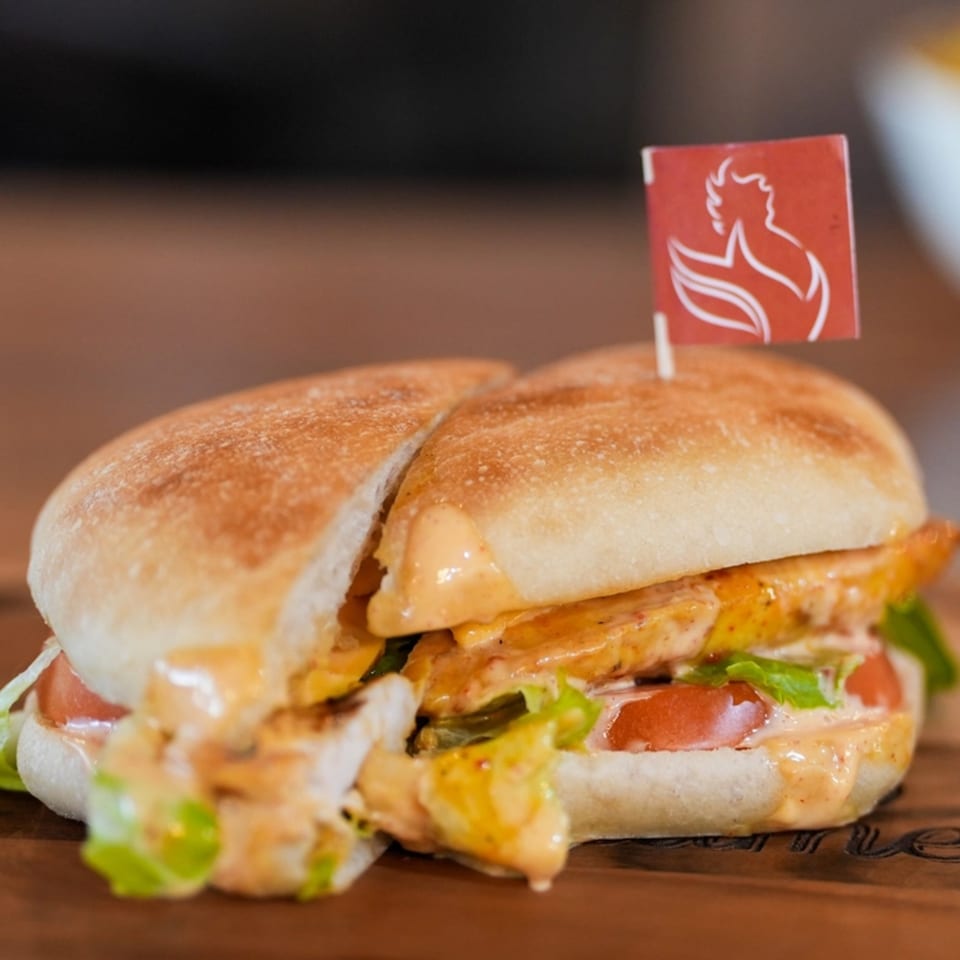 A peri-peri chicken sandwich, cut in half, with lettuce, tomato, and sauce on a wooden board.